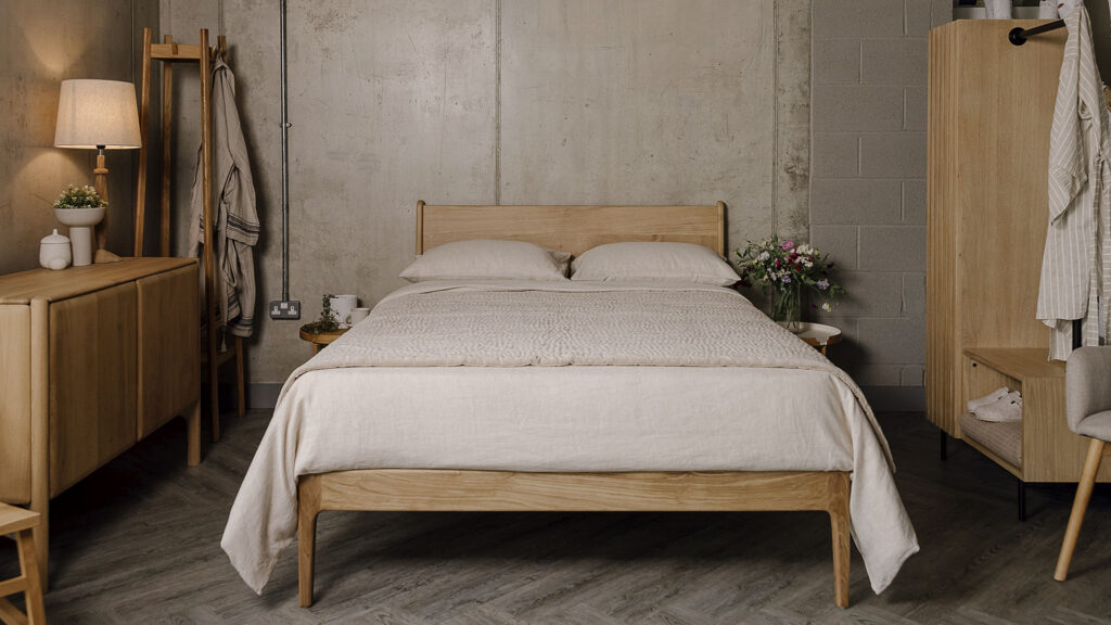Zoomed out shot of the English oak Camden, showing its headboard and foot-rail in full view. It is dressed in the organic ecru linen bedding and a soft beige stitched velvet bedspread. There are two round-top bedside tables on both sides of the bed. The Ethnicraft Pi sideboard in oak and the Tower Coat stand sit to the left of the bed base and the Modern oak Open Wardrobe sits to the right side of the bed.