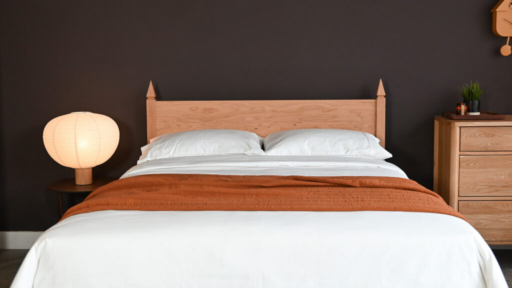 Cherry Samarkand bed base with white cotton bedding and a terracotta bedspread. Next to an oiled oak chest of drawers to the right and an oak lamp with a large paper shade to the left.