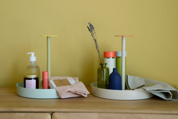 2 Colourful round trays with handles. One light green with yellow topped handle carrying hand soup, a candle and other bathrooms supplies. The other grey with orange handle carrying a vase and flower, a cloth and a colourful salt and pepper grinder.