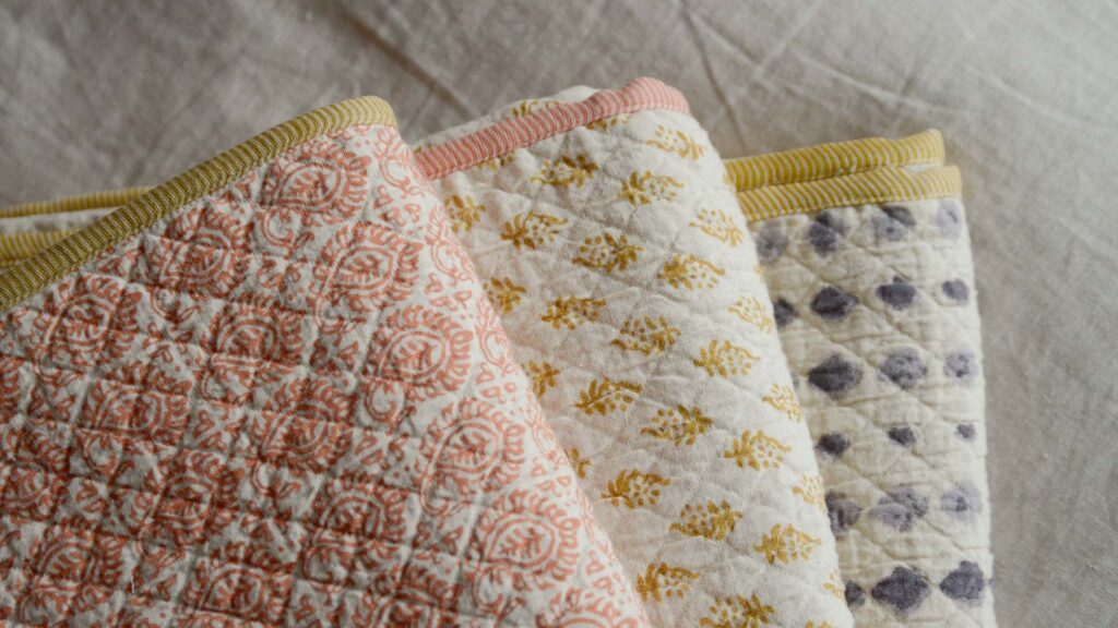 3 bedspreads neatly folded and stacked in a fan. Terracotta tiles, mustard diamonds and purple ink spots.