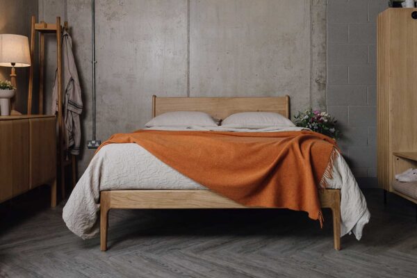 Front view of a bedroom scene with an English oak bed in the centre, surrounded by an oak sideboard, a coat stand with robe hanging on it and an open rail and shelf set. Bed made with ecru linen and a beige textured bedspread, with a burnt orange lambswool throw draped over the top.