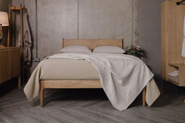 Zoomed-out shot of the English oak Camden, showing its headboard and foot-rail. It is dressed with the floral textured bedspreads in the colours ecru and sand. On one side of the bed sits the Ethnicraft oak Pi sideboard and the Tower oak coat-stand, and on the other side of the bed sits the Modern Oak Open Wardrobe.
