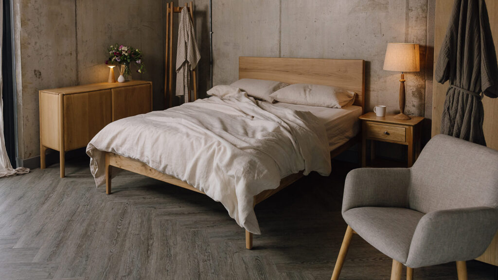 3/4 angle image of oak bed with large wooden headboard. Bed dressed in rumpled linen, other oak furniture in the room scene.