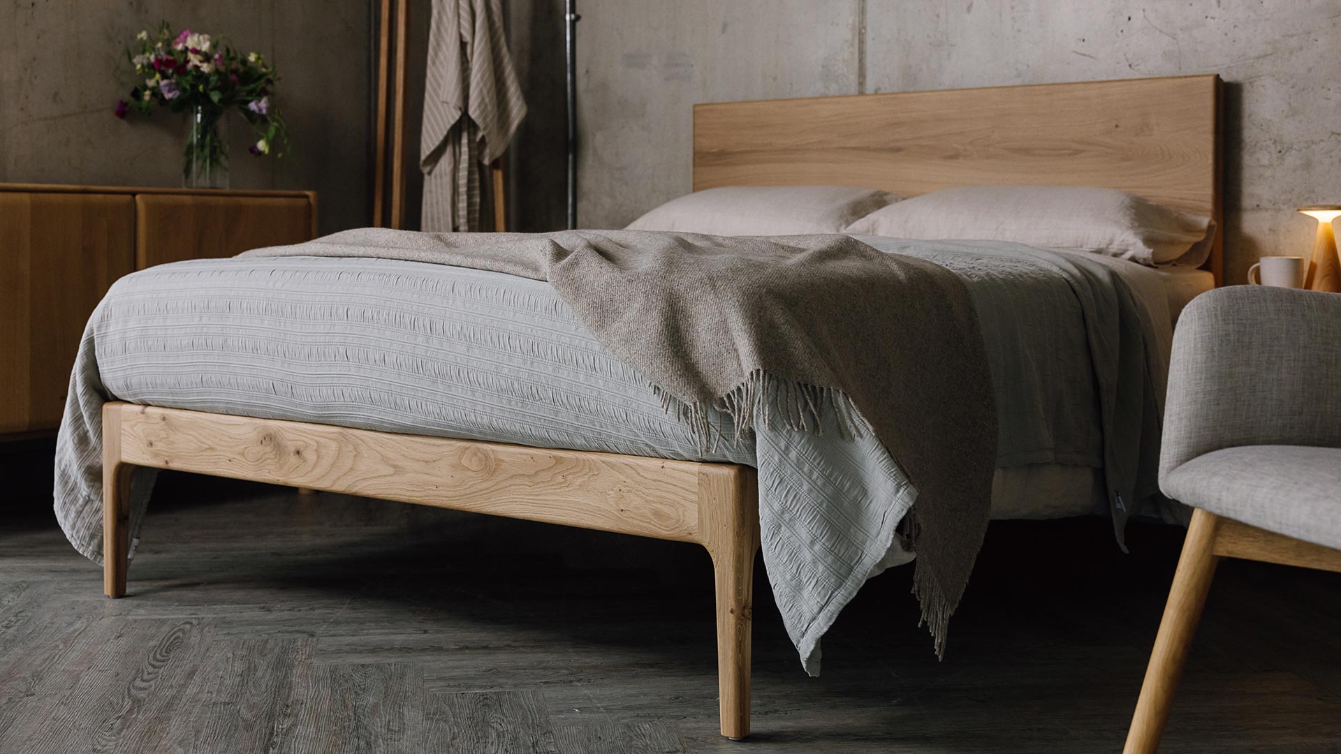 A low angled shot of the English oak Chiswick, focusing on the foot-rail section of the bed. The bed is dressed in the organic linen ecru bedding, a silver grey embroidered bedspread and a beige pure wool throw.