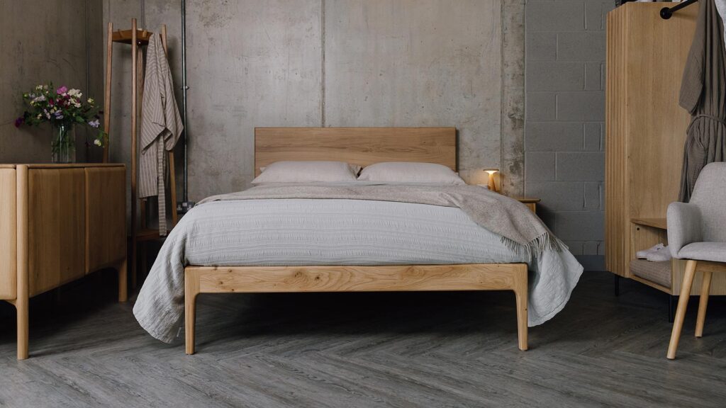 A forward-facing shot of the English oak Chiswick. It is dressed in the organic linen bedding in ecru, which is covered by an embroidered striped bedspread in silver-grey and a pure wool beige throw.
