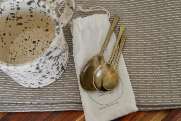 Forged iron spoons sat on the bag that they come packaged in, to show how they will arrive to you. Pictured also is a grey and white mug to give a sense of setting.