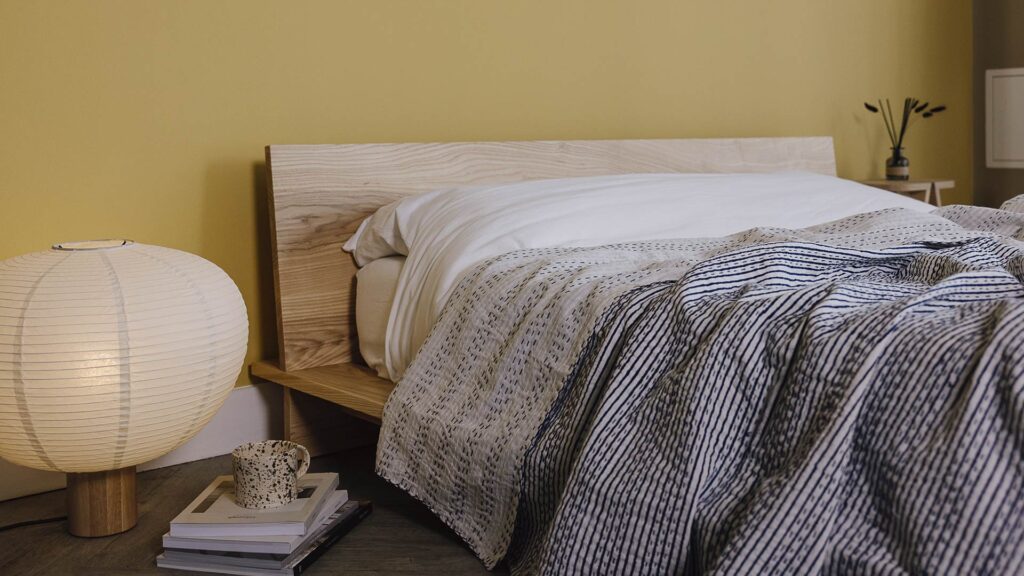 Low platform bed dressed in white linen with a blue striped bedspread laid over the top. A stack of books, speckled mug and Japanese inspired large lamp on the floor to the side.