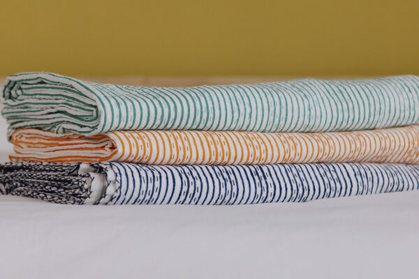 3 striped kantha bedspreads neatly folded and stacked on a white sheet. Top to bottom - green, orange and dark blue.