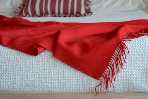 Image showing very bright red alpaca wool throw on the low Oregon bed base. The bed has a textured white bedspread on it that the throw is draped over. You can also see a red and white striped cushion in the background.