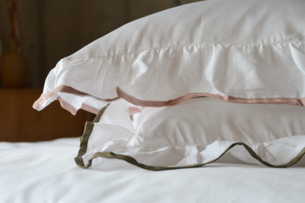 A stack of two pure white pillowcases with ruffles edges. Once with a dusty pink edge, the other olive green.