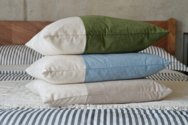 A stack of Velvet Block Cushions in all three colour options. The top cushion is an olive green velvet, the middle is a pale blue velvet and the bottom is a warm stone velvet. Each cushion is also made partly of linen, which can be seen to the right hand side. The cushions are sat on a walnut Dalston bed with black and white bedding.