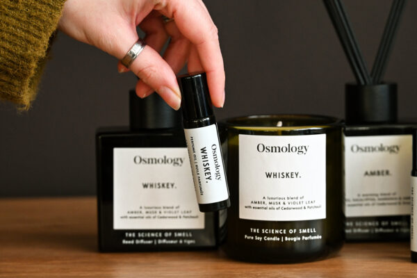 A close up image of a hand holding up the Aery Osmology roll-on perfume oil in the 'Whiskey' scent. In the background, the Aery diffusers, roll-on perfume oils and candles are visible in the Whiskey and Amber scents.