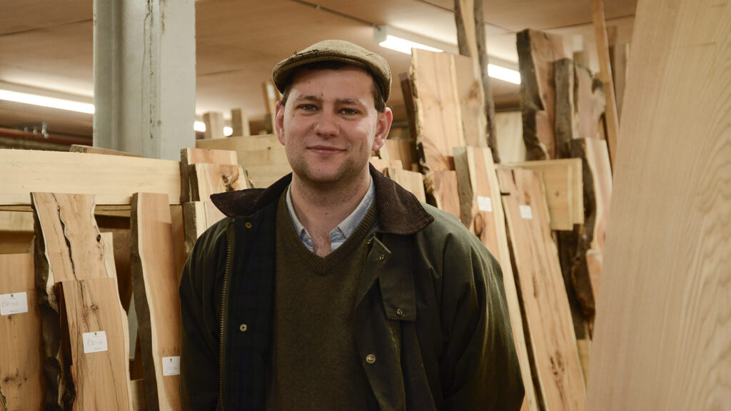 Image of a man smiling. He is wearing a flat-cap and a jacket. Behind the man, there are stacks of raw timber. The man is the focal point of the image.