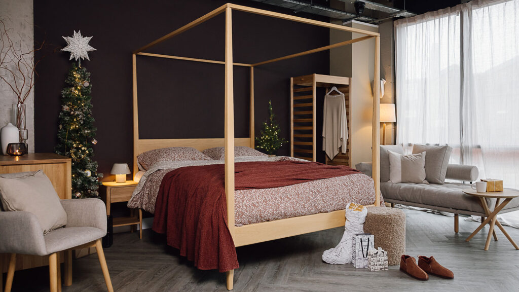 Bedroom scene with Christmas trees and gifts at the foot of the bed. Bed dressed in red linen, oak furniture and a grey lounger also featured.