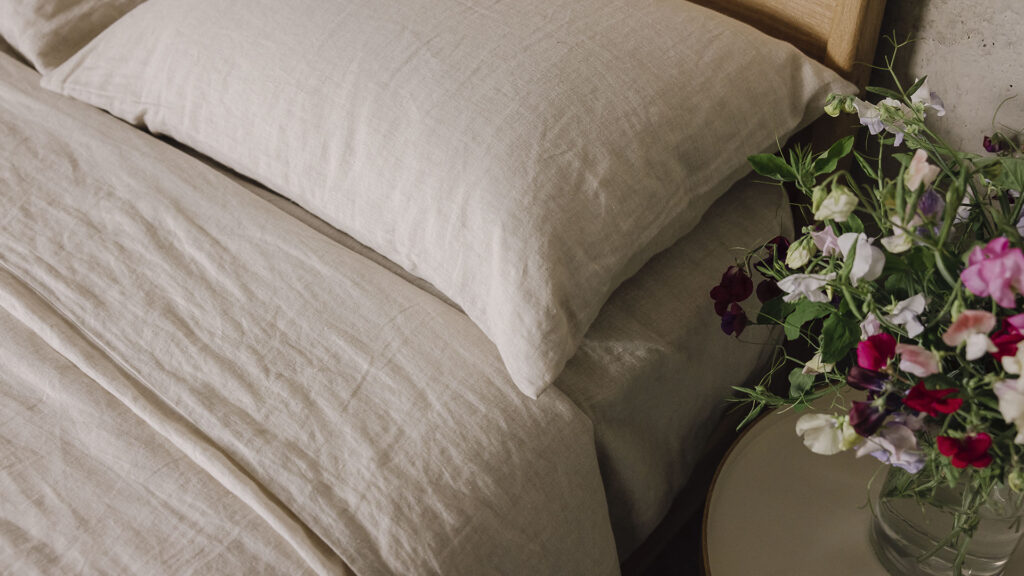 Close up shot of a pillow and duvet cover dressed in the organic ecru linen bedding. There is a table next to the bed which has a clear vase filled with colourful flowers.