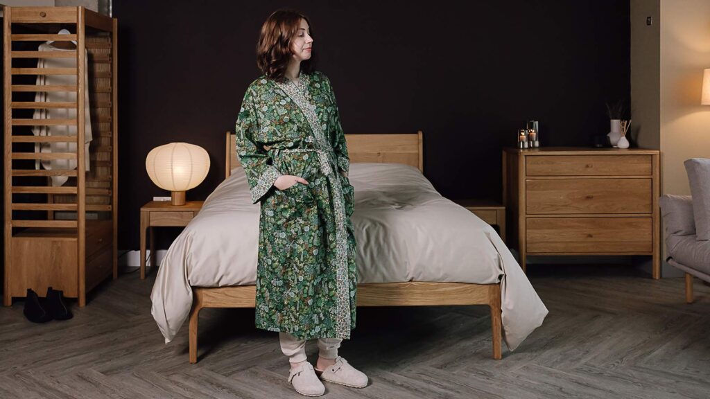 Model in floral green liberty print robe stood in front of an English oak bed frame.