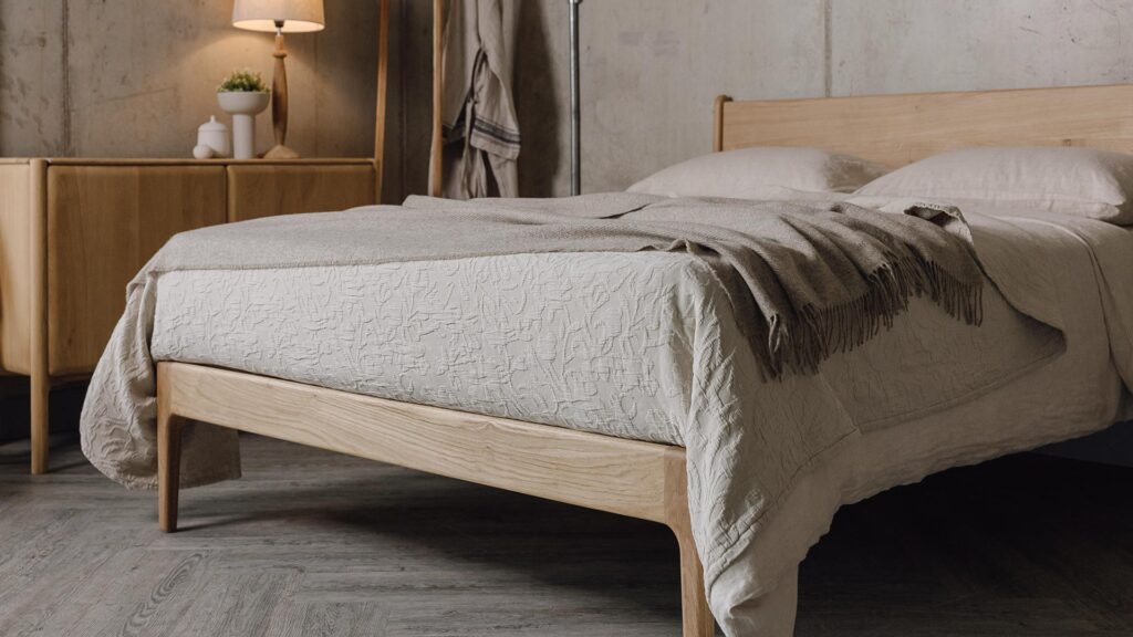 3/4 angle photo of an oak bed with stone linen and a greige lambswool throw.