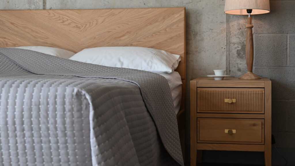 Image shows the grey colour option for the modern quilted bedspread. The item is shown dressed on the Oak Chiswick bed with an oak bedside table, lamp and porcelain cup to the right of the image.