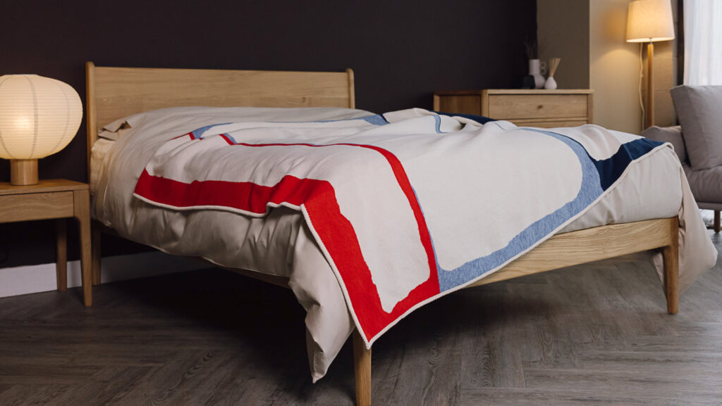 Oak bed with light taupe bedding. Blue and red blanket draped over the top. Other oak furniture also in shot.