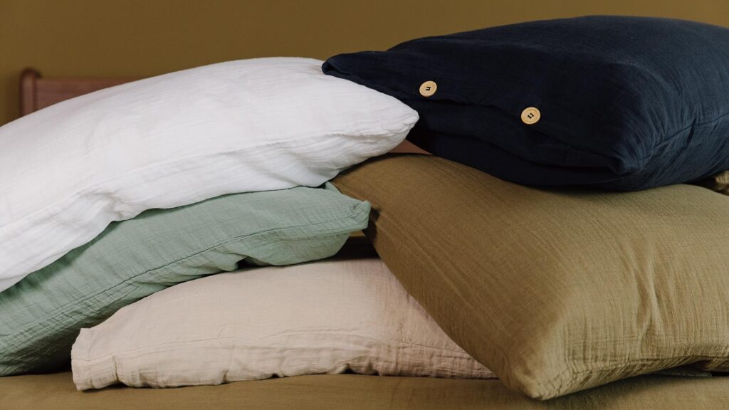 A stack of pillows dressed in muslin pillowcases, showing the colours white, eucalyptus, deep navy, khaki and natural.
