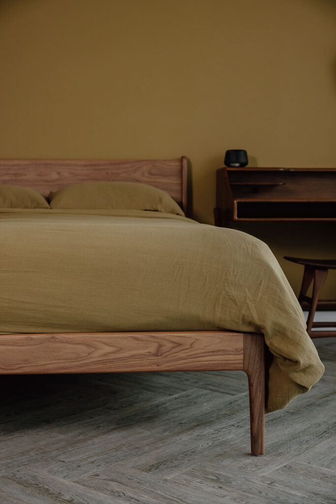 Portrait image of the Camden bed base in solid walnut, dressed in khaki green muslin bedding