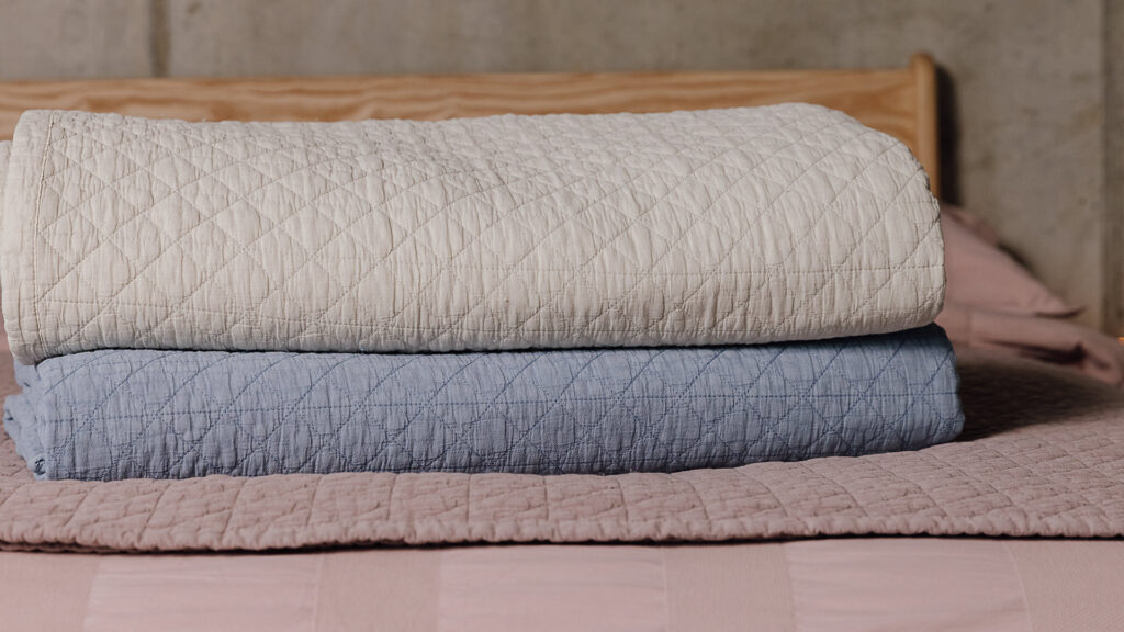 Stack of three quilted bedspreads. From top to bottom: ecru, blue, pink.