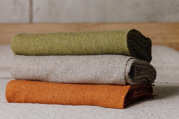Stack of three lambswool merino throws on a bed. Top to bottom - Green, griege, burnt orange.