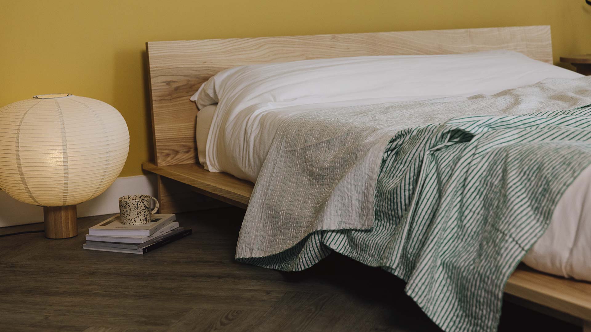 close up of ash bed frame with white linen. Green striped bedspread laid over the top. Stack of books, Japanese style paper lamb and speckled mug to the side.