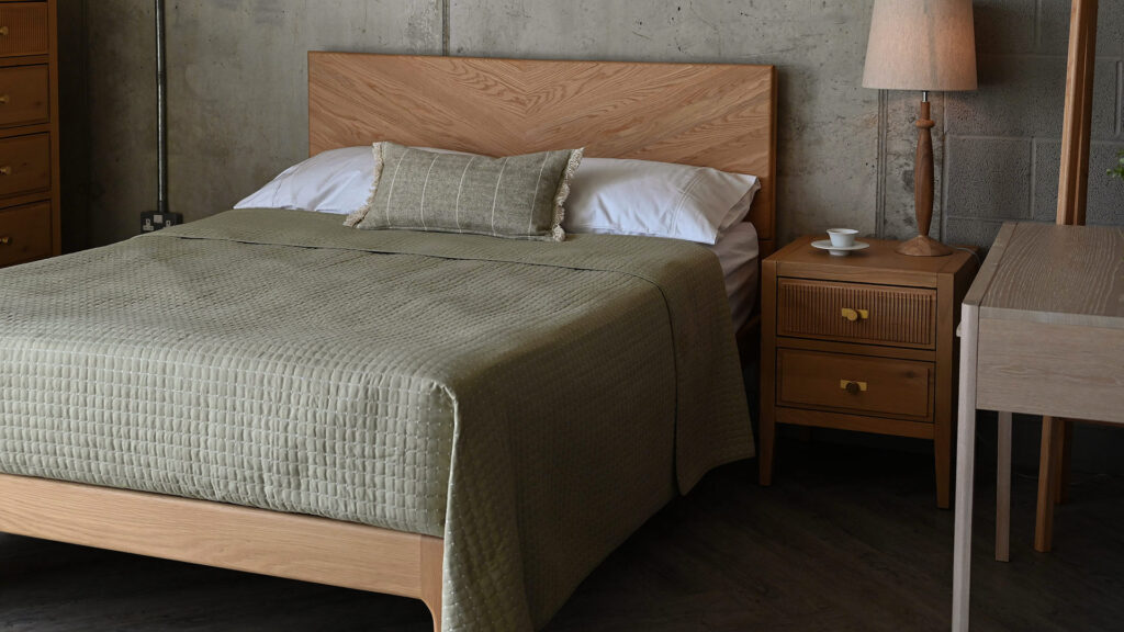Sage Green modern quilted bedspread with a matching sage green breakfast cushion. The bedding is photographed on the Chiswick bed made in solid oak, with an oak bedside table and pale oak dressing table to the right of the image.