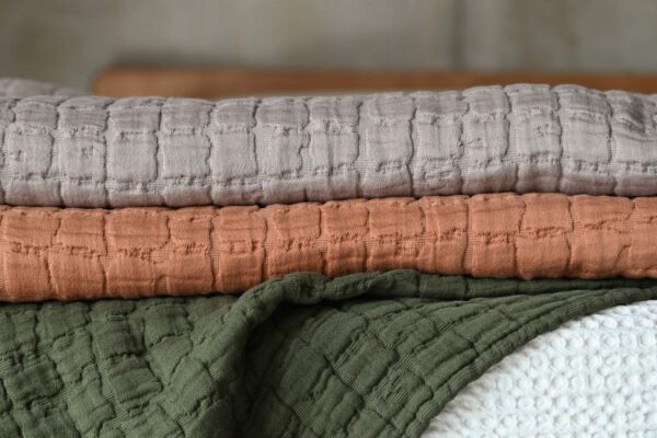The taupe and the terracotta textured bedspreads folded on top of an unfolded deep olive bedspread.