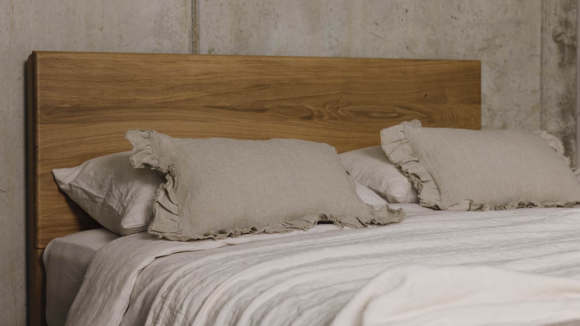 close up of the top of a bed. Oak headboard with grain detail, neutral bedding and small ruffled edge linen cushions.