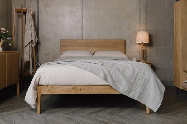 A forward facing shot of the English oak Chiswick, dressed in the organic linen bedding in ecru. The bedding is mostly covered up by a embroidered striped bedspread in the colour silver-grey, which also overhangs the bottom right-hand corner of the bed base.