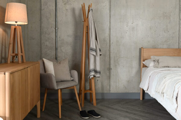 Spire hat and coat stand in modern room setting. The coat stand has a cotton robe hanging from it, and stands next to a grey upholstered chair with a cushion on it, as well as a modern oak sideboard. The image also features the Camden bed base in English Oak to the right hand side of the image.