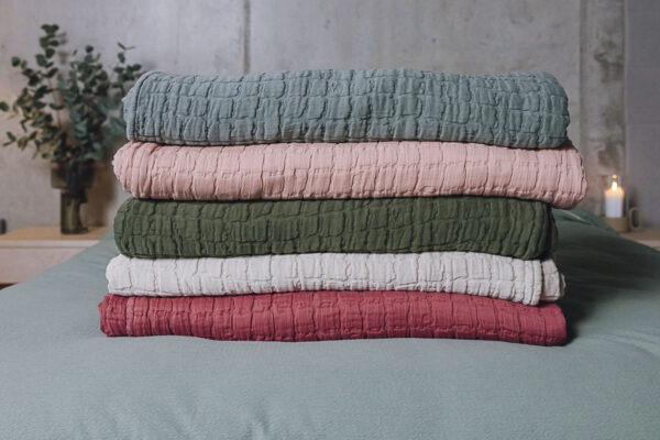 Stack of 5 textured bedspreads neatly folded on a bed. From bottom to top, raspberry, light grey, dark green, soft pink and eucalyptus.