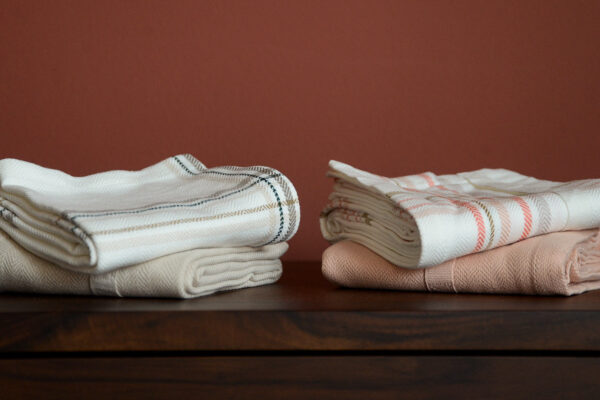 Stack of 2 folded stone tea towels and a stack of 2 folded blush tea towels.