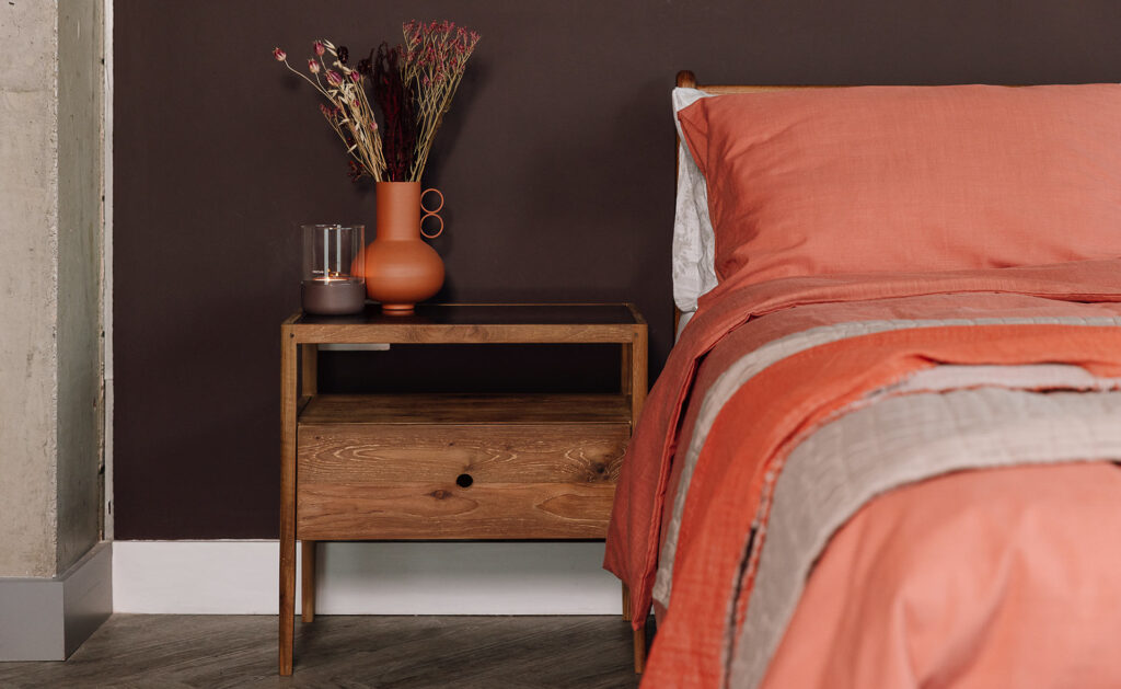 A mid-century style bedside table; the Spindle Table in reclaimed teak. A vase of flowers sits atop the table in a terracotta colour, with a lit candle placed next to it. The bedside table features a large drawer with a small hole in the front of it for a handle.