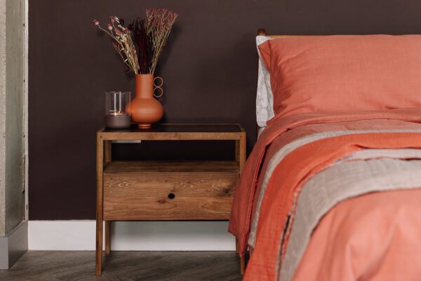 A mid-century style bedside table; the Spindle Table in reclaimed teak. A vase of flowers sits atop the table in a terracotta colour, with a lit candle placed next to it. The bedside table features a large drawer with a small hole in the front of it for a handle.