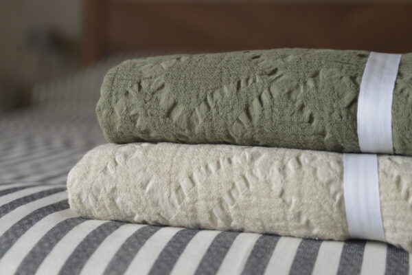 Stack of folded textured cotton throws, showing olive green on top of light ecru, laid on top of black and white stripe bed linen.