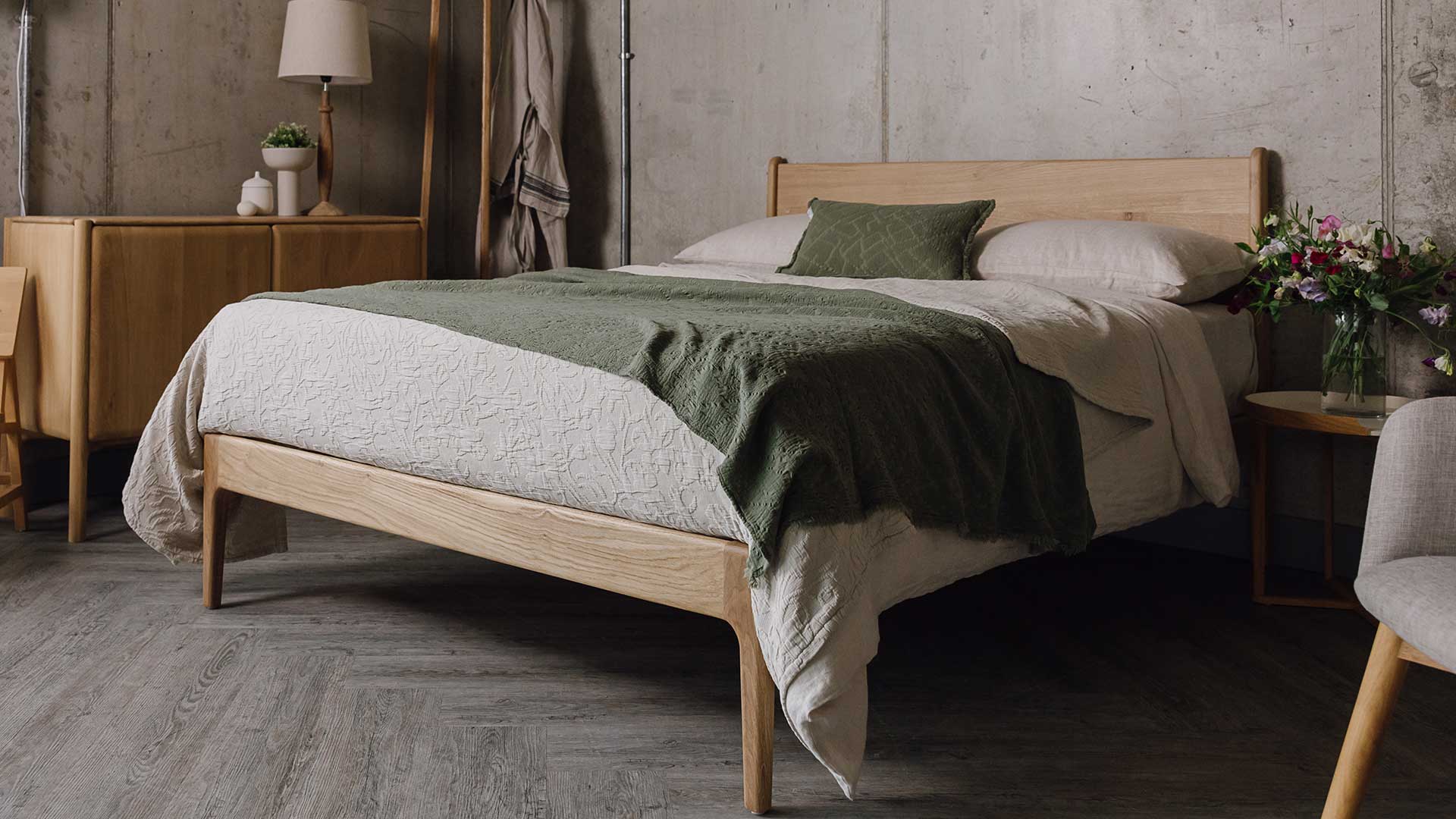 English oak bed, headboard and footboard rail visible. Bed dressed in ecru bedding, with an olive green pillow, and an olive green textured throw draped on the top. Bed room scene also featuring an oak sideboard, a coat stand, a circlular table and a grey chair.