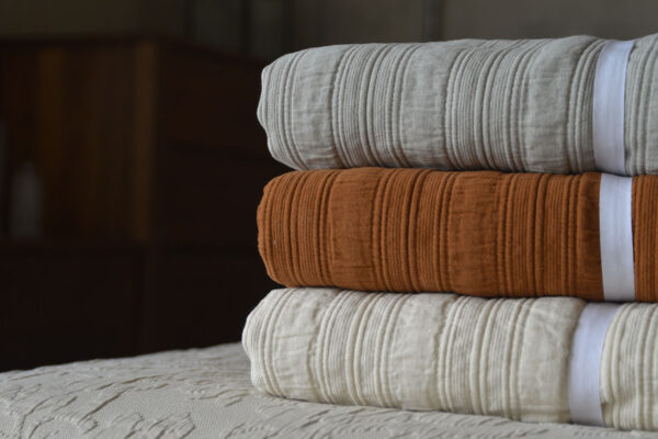 Stack of folded stripe cotton bedspreads, in the colours silver grey (top), cinnamon (middle) and ivory (bottom).