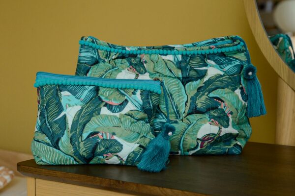 Two blue and green tropical print toiletry bag with large tassels, one small, one large, sat on dark table top with mirror in the background.