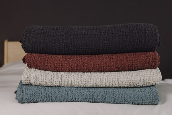 Stack of 4 waffle textured bedspreads neatly folded on a bed. From bottom to top, black, red, ecru and green.