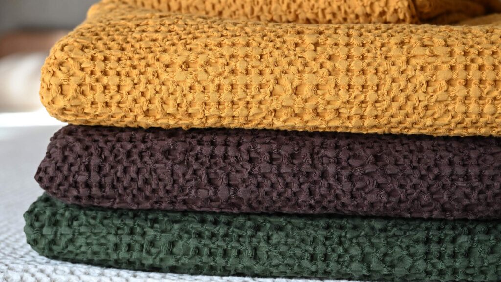 A close up of a folded stack of waffle textured bedspreads, showing (from top to bottom) colours Mustard, Espresso and Deep Olive.