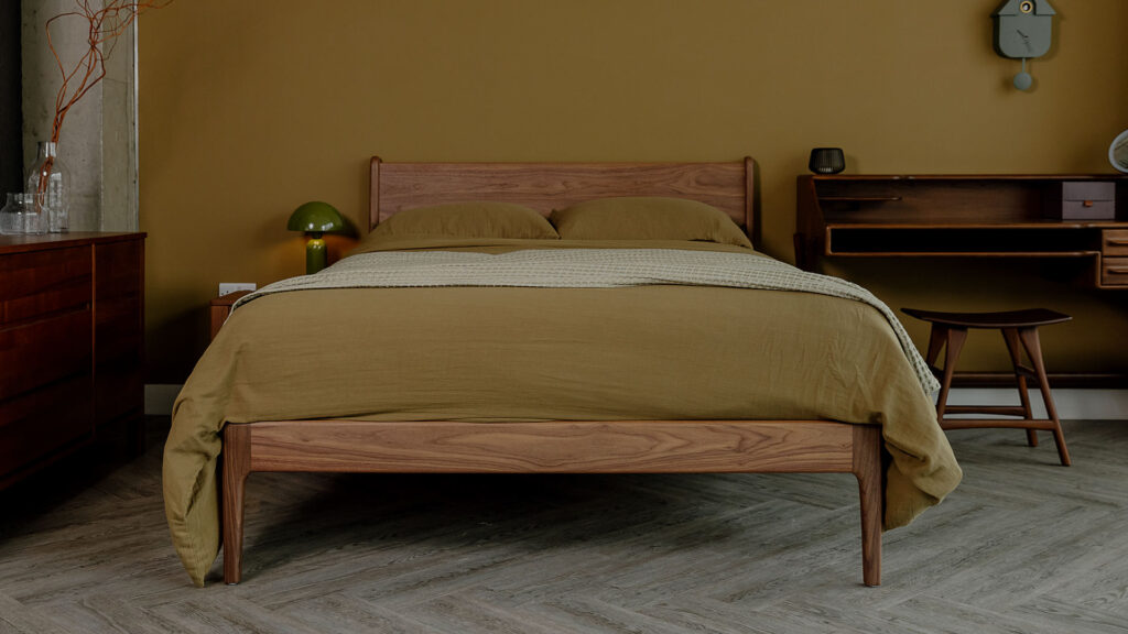 A walnut Camden bed dressed in khaki muslin cotton bedding and a sage green waffle throw. Shown next to the bed is an Ethnicraft Flow Desk and Osso Stool, both in solid brown teak.