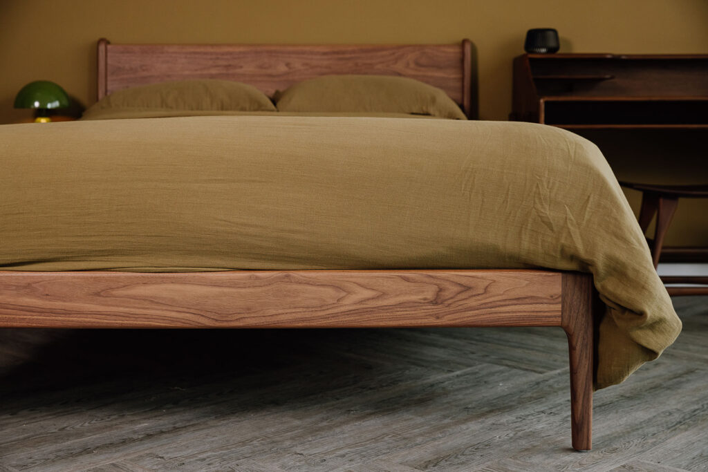 Close up of a walnut Camden foot rail and leg. The bed is dressed in plain khaki muslin bedding.