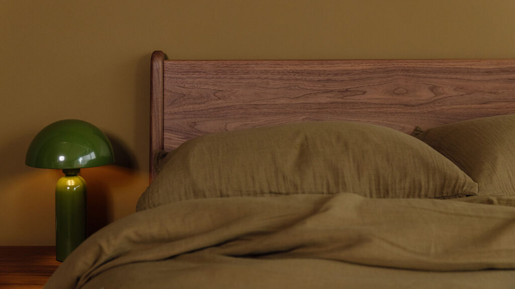 close up of walnut headboard on a Camden bed. Green lamp to the side. Bed dressed in olive muslin.