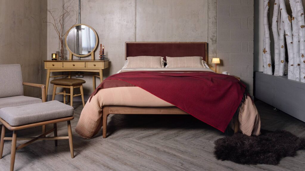Walnut Carlisle bed frame with dark red headboard with red ben linens. Dressing table, a grey chair and stool and a dark faux fur rug also in the photograph.